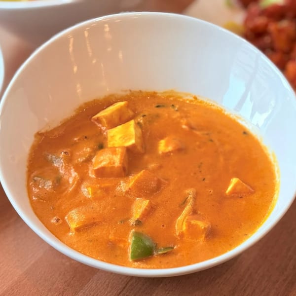 A white bowl filled with Amrut's Indian paneer butter masala, featuring paneer cubes and green bell peppers.
