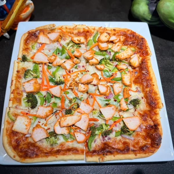 Square Indian pizza topped with chicken, broccoli, bell peppers, and shredded carrots on a white plate.
